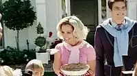Image of a blond woman. She is a housewife and carries a pie in her left hand. Her hair is styled in 1950s fashion. She is wearing a pink polo shirt. Surrounding her are her husband and children.