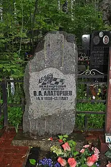 A grave of dark stone behind a bed of pink and blue flowers and in front of dark green foliage. The name on the stone reads "V. A. Alatortsev" in Cyrillic letters.