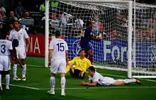 Four men in white shirts: three are standing and one is lying on the ground. A man wearing a yellow shirt is sitting on the ground. A man in a dark shirt is picking a football out of a goal.