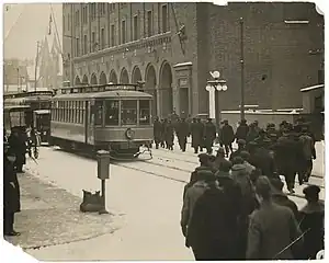 A black and white photograph of a group of people and a streetcar outside of a brick building