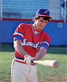 A man in a red baseball jersey with "Sounds" written on the front in white and blue and a blue cap with a white "N" on the center stands on a baseball field swinging a bat.