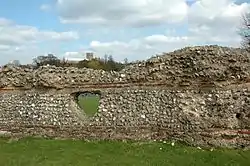 Mortared wall with stacked thick stone layers over thin red brick layers, with a triangular tunnel through