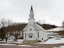 A small, steepled wooden church and wooden house in front of a hillside cemetery