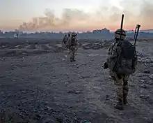 Colour photograph of marines patrolling in single file over rough, almost barren, terrain with buildings on the horizon.