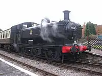 A black pannier tank locomotive is passing along a single track through woodland. The locomotive is pulling a train of a covered red wagon, three grey open wagons, and a red guard's van.