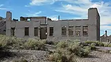 A roofless two-story masonry building rests in a setting of low shrubs and gravel under a partly cloudy sky. The building has a wood-framed doorway but no door, and many window openings but no glass. Most of the upper walls have collapsed and several cracks appear in the remaining walls. A hill rises to the left, and other hills and ruins are visible in the distance to the right.