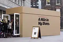 Shipping container decorated to look like a giant shoebox, with a queue of people at its doors.