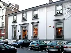 A two-storey white building with grey window frames, several cars are parked in the foreground.