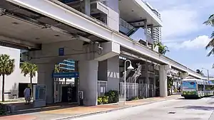 An elevated metro station viewed from ground level, with a bus driving on a street in the foreground.