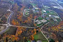 An aerial view of a school campus in a wooded area. The trees around the campus have red leaves. Roads cut through the middle of the wood toward the school.