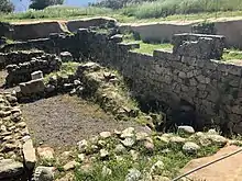 Pieces of rough stones form a rectangular blank space in the lower left hand side of the image. It is encircled by a taller stone wall on the right, and the entire area is covered in overgrown weeds.