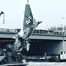 A severed airplane tail section hangs from a crane just above the water, guyed by crew on barges. A low, steel beam bridge with granite block piers stands behind, it's railing lined with onlookers.