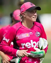 Young woman with a short blonde ponytail wearing a dark blue T-shirt, baseball cap and trackpants with gold stripes. Advertising logos of Adidas and Commonwealth Bank are present on the clothes. She is smiling broadly.