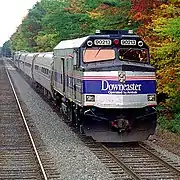 A depowered diesel locomotive with gray paint and a black roof. A wide blue stripe and two thinner red stripes are on the sides and front.