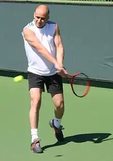 An older, bald man plays tennis. He's wearing a white sleeveless shirt and black shorts.  He is bald and is holding a red tennis racket.