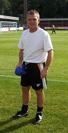 A middle-aged man, wearing a white T-shirt and black shorts, standing on a grass field.