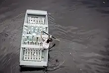 Overhead view of a young girl, alone, rowing through  murky floodwaters on a wooden boat carrying several large plastic water bottles