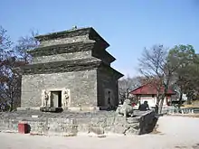 A huge three-story pagoda stands against blue skies. The pagoda is made with bricks of dark gray stone. Colorful lanterns are lined up.