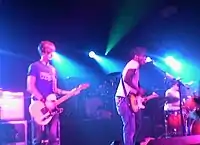 Two guitarists and a drummer are performing a song live on a stage lit by blue concert lights