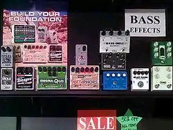 A music store display of effect pedals for bass is shown. The pedals have foot-operated switches to turn the effect on an off and knobs for controlling the tone.