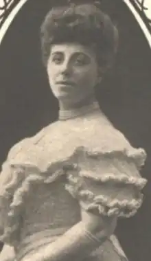 A white woman wearing her hair in an updo, and a light-colored gown with heave ruffles on the short sleeves and chest. She is wearing elbow-length gloves.