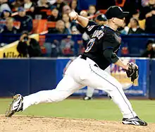 A man wearing a black baseball jersey and cap and white baseball pants throwing a baseball with his left hand