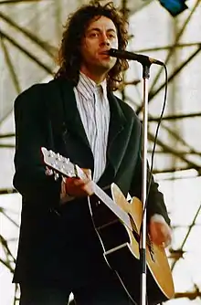 Bob Geldof, a Caucasian man in his mid-thirties, is on stage, singing into a microphone and playing a left-handed acoustic guitar. He wears a white shirt and a dark green jacket.