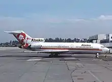 Right side view of an aircraft parked on the tarmac, with a tree-covered hill as well as some clouds and buildings in the background