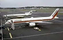 Side view of twin-engine jet on tarmac, with attached airstairs and support vehicle, along with a trijet aircraft in the background.