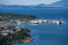 The waterfront on the left side with multiple large ships and small sail boats in the water. Mountains are visible in the distance