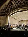  In a ivory concert hall, a stage full of classical musicians and conductor are standing at the end of a performance