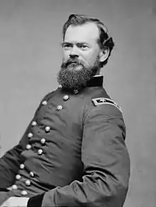 Black and white photo of a bearded man sitting and leaning backward. He wears a dark military uniform with two rows of buttons and the two stars of a major general on the shoulder tabs.