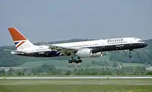 Side view of aircraft in flight with extended gear, against a grassy hill backdrop