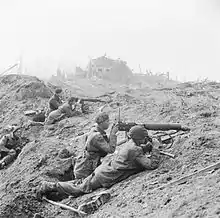 A black and white photograph of a battle scene in which two Vickers machine guns are firing from behind cover of a pile of rubble. Each machine gun is crewed by two men, a firer and an observer with a pair of binoculars. Another soldier is looking on from behind the crews