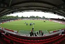 The inside of a football ground as photographed from one of the touchlines. Red seating is visible in the foreground, with the pitch in the middle and the opposite touchline in the background. A standing-only area is visible to the right of the picture. Football players and officials can be seen on the pitch.