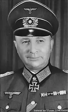 The head of an elderly man, shown from the front. He wears a peaked cap and a military uniform. His facial expression is a determined and confident smile; his eyes looks to the right of the camera.