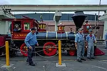 A red steam locomotive with a 4-4-0 wheel arrangement (four leading wheels, four driving wheels, and no trailing wheels) and its tender