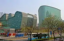 Two irregularly shaped greenish glass-faced buildings rise over a street lined with freshly budding trees on which a blue and white trolleybus is stopped