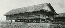 Black-and-white image a grass nipa hut raised a few feet off the ground by wooden supports. Another hut can be seen in the background.