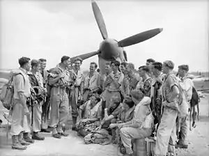 Full-length portrait of moustachioed man in flying suit with parachute holding the attention of a group of twenty or so similarly kitted men in front of a single-engined military monoplane