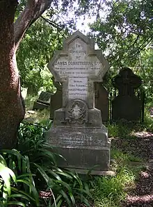 A large stone headstone sits next to the trunk of a large and leafy tree, with other gravestones visible in the background. The stone is weathered and mossy. An inscription bears the names of James Donnithorne and Eliza Emily Donnithorne.