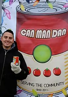 Johnstone standing in front of a large poster of a can. The can reads, "Can Man Dan. Serving the community. Since 2011."