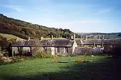 A green field with rows of two-storey buildings with tall chimneys beyond and wooded hills behind under a blue sky