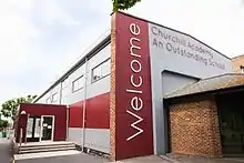 The entrance to Churchill Academ school with a 'welcome' sign and the school logo attached to the wall