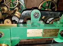 Close-up colour photograph of the clockwork installed in the tower. The metal body is painted green with a label affixed to the front that reads as follows: "Overhauled by Bristol Goldsmiths' Alliance 1939." To the right of this label is a brass plaque with the following inscription: "Repaired by James R. Cornish of Langford 1975."