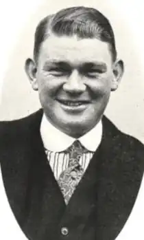 A smiling white man wearing a dark suit, striped shirt, print tie, and round white collar; he is cleanshaven and has short side-parted hair