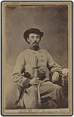 Sepia photo shows a seated man with a moustache wearing a black hat and a light gray military uniform.