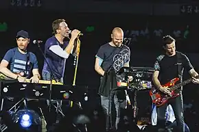 Four men perform on the stage, from left to right, they are on the piano, microphone, drum pad and bass