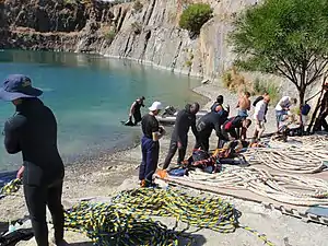 A group of about 12 divers on the shore of a flooded quarry preparing surface-supplied diving equipment for diver training exercises. Several umbilicals are laid out for use in figure 8 coils.