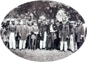 A photograph showing a large group men, most of whom wear uniforms, standing beneath shade trees